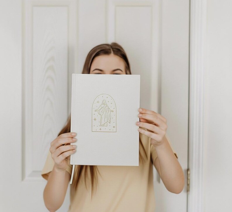 Pu&ograve; includere: Un diario color crema con un'illustrazione dorata di una figura all'interno di una cornice ad arco, circondata da stelle e punti. Il diario ha un segnalibro a nastro abbinato. La copertina &egrave; liscia e il design &egrave; minimalista.