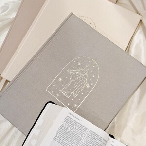 Pu&ograve; includere: Una pila di libri con copertine dai toni neutri, tra cui un libro grigio chiaro con un'illustrazione dorata di una figura all'interno di un arco e stelle. Un libro aperto con bordi neri e testo in primo piano. Il tema &egrave; religioso.
