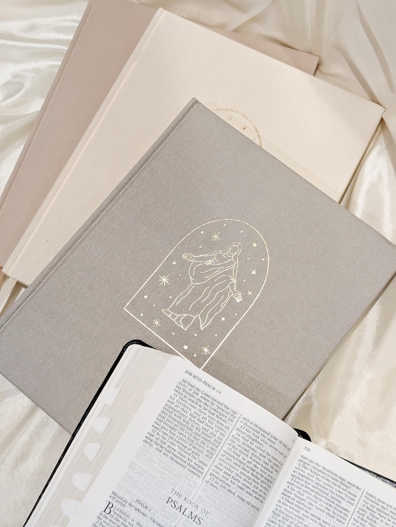 Pu&ograve; includere: Una pila di libri con copertine dai toni neutri, tra cui un libro grigio chiaro con un'illustrazione dorata di una figura all'interno di un arco e stelle. Un libro aperto con bordi neri e testo in primo piano. Il tema &egrave; religioso.