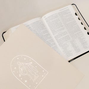 Pu&ograve; includere: Una Bibbia aperta con bordi neri e una copertina color crema. La copertina presenta un disegno a tratto bianco di una figura all'interno di una cornice ad arco, circondata da stelle. Le pagine della Bibbia sono piene di testo.
