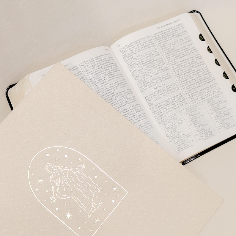 Pu&ograve; includere: Una Bibbia aperta con bordi neri e una copertina color crema. La copertina presenta un disegno a tratto bianco di una figura all'interno di una cornice ad arco, circondata da stelle. Le pagine della Bibbia sono piene di testo.