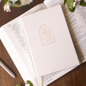 The image shows a beige christus notebook resting on top of a bible. The front cover features a illustration of Jesus Christ with outstretched arms. It is a religious Bible journal. The journal is neutral and textured.