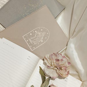Puede incluir: Un conjunto de diarios en tonos neutros, con un acento floral. Los diarios tienen un diseño celestial en la portada. Un diario está abierto, revelando páginas rayadas con los días de la semana impresos en la parte superior.