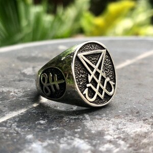 May include: A silver ring with a black sigil of a pentagram with an upside-down cross inside. The ring has a textured surface and a raised edge.