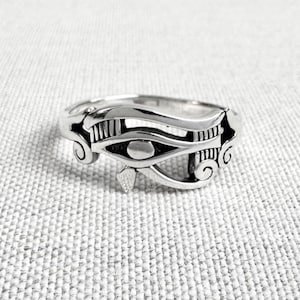 May include: A silver ring with an Eye of Horus design. The ring has detailed features, including the eye, eyebrow, and markings. The ring is set against a textured, light grey background.
