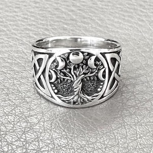 May include: A silver ring featuring a detailed design of a tree with crescent moons and Celtic knotwork. The ring has a wide band and a polished finish, showcasing the intricate details of the celestial and nature-inspired imagery.