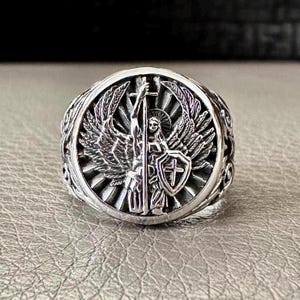 May include: Silver ring featuring an angel design. The angel is holding a sword and shield with a cross on it. The ring has an intricate design with a sunburst pattern.