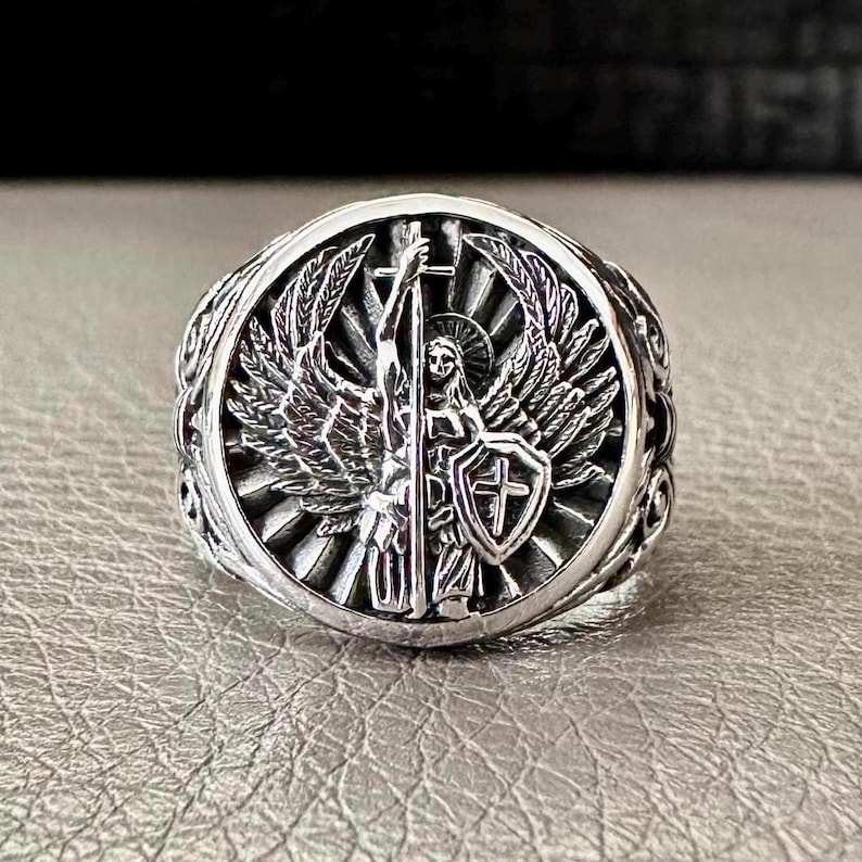 May include: Silver ring featuring an angel design. The angel is holding a sword and shield with a cross on it. The ring has an intricate design with a sunburst pattern.