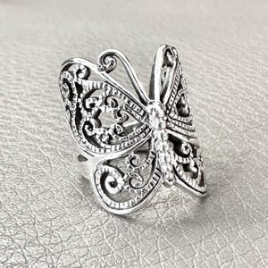 Puede incluir: Un anillo de mariposa plateado con intrincados detalles de filigrana. El anillo presenta un diseño de mariposa detallado con alas caladas y un cuerpo texturizado. El anillo está sobre un fondo gris claro.