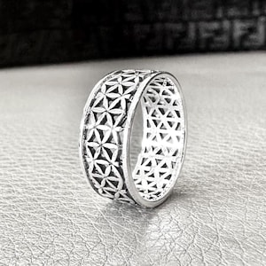 Pode incluir: Um anel de prata com um padrão floral geométrico. O anel apresenta um design repetitivo de formas interconectadas, criando um efeito vazado. O anel é exibido em uma superfície reflexiva.