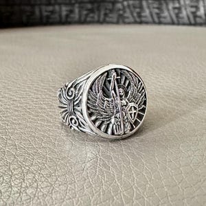 May include: A silver ring with an ornate design featuring an angel with wings holding a sword and shield.