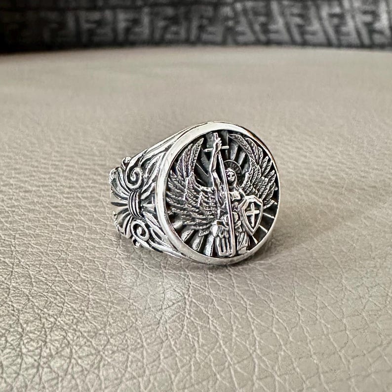 May include: A silver ring with an ornate design featuring an angel with wings holding a sword and shield.