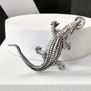 Könnte beinhalten: Detaillierter silberner Alligator-Anhänger mit strukturierten Schuppen und einem Ring oben. Der Anhänger ist eine realistische Darstellung eines Alligators mit sichtbaren Krallen und einem langen Schwanz.