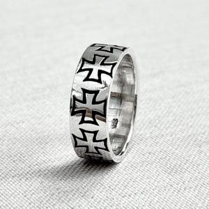 Könnte beinhalten: Ein silberner Ring mit einem sich wiederholenden schwarzen Malteserkreuzmuster. Der Ring besteht aus glänzendem Metall mit einer glatten, polierten Oberfläche. Das kühne Design ist für den Alltag geeignet und hat ein auffälliges Aussehen.