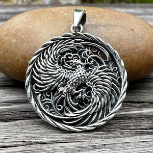 May include: A detailed silver pendant featuring a phoenix design. The pendant showcases the mythical bird with outstretched wings, surrounded by intricate scrollwork. The pendant is round and has a bail for a chain.