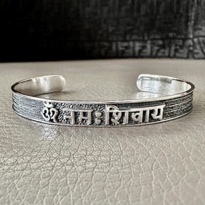Puede incluir: Una pulsera de plata con la frase sánscrita "Om Namah Shivaya" grabada.