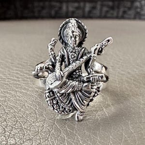 Op de afbeelding: Een zilveren ring met een gedetailleerd beeldhouwwerk van een godheid die een snaarinstrument bespeelt. De godheid is afgebeeld in zittende positie met ingewikkelde kleding en sieraden.