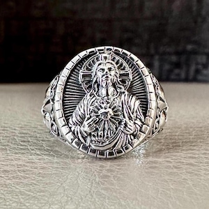 Pode incluir: Um anel de prata com um design detalhado de Jesus Cristo segurando um coração. O anel é ornamentado e apresenta um fundo texturizado.
