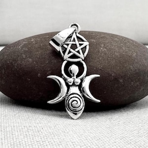 May include: A silver pendant featuring a stylized depiction of a goddess figure with crescent moons and a spiral design. Above the figure is a five-pointed star within a circle. The pendant is displayed on a dark, textured stone.