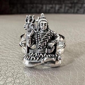 May include: Silver ring featuring a detailed depiction of the Hindu deity Shiva, seated with his trident, in a meditative pose.