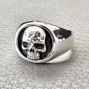 May include: A silver skull ring with a detailed skull design set in a circular frame. The ring has a polished finish and a wide band, suitable for a bold statement piece.