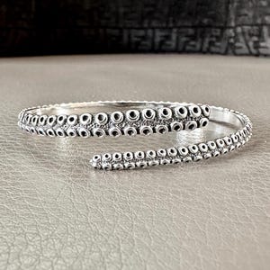 May include: A silver-colored cuff bracelet with a tentacle design. The bracelet wraps around the wrist with a detailed pattern of small, circular suckers. The open-ended design allows for easy wear and adjustment. The bracelet is made of a shiny, metallic material.