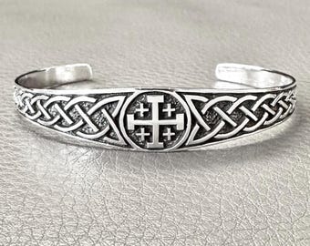 May include: A silver cuff bracelet featuring a Jerusalem cross design at the center, surrounded by intricate Celtic knot patterns. The open-ended bracelet is made of polished metal, reflecting light. The bracelet is a piece of jewelry.