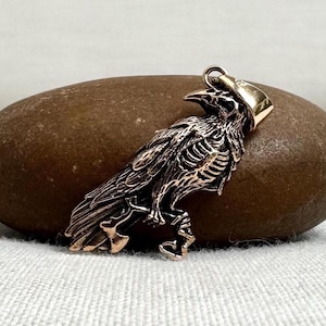 May include: A metallic bronze-toned pendant shaped like a skeletal raven in profile. The bird figure features detailed rib cage and wing feather textures, with a smooth, polished metal bail attached at the top for hanging on a chain or cord.
