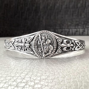 Könnte beinhalten: Silberarmband mit einem keltischen Knotendesign und einem runden Medaillon, das den Heiligen Christophorus zeigt, der ein Kind trägt. Das Medaillon ist mit "Saint Christopher" beschriftet.