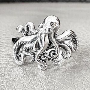 Könnte beinhalten: Ein silberner Oktopus-Ring. Der Ring zeigt ein detailliertes Oktopus-Design mit acht Tentakeln und einer strukturierten Oberfläche. Der Oktopus ist der Mittelpunkt, mit seinem Kopf und seinen Tentakeln deutlich sichtbar. Der Ring ist wahrscheinlich aus Sterlingsilber gefertigt.