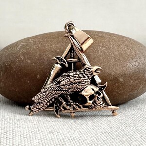 May include: Bronze-colored pendant featuring a raven perched atop a skull, set within a triangle. Swords and a hammer are crossed behind the raven. The pendant has a loop for a chain.
