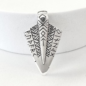 May include: Silver pendant with a triangular shape. It has etched geometric patterns and runic text. The top has a loop for a chain, and the bottom is pointed. The pendant is likely made of metal, possibly silver or a silver alloy.