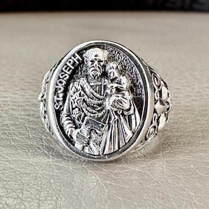 May include: A silver ring with a detailed design featuring Saint Joseph holding a baby Jesus. The ring has a textured band with a circular design.