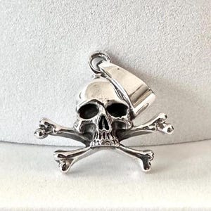 May include: Silver skull and crossbones pendant charm. The skull is detailed with eye sockets and a nose. The crossbones are simple and symmetrical.