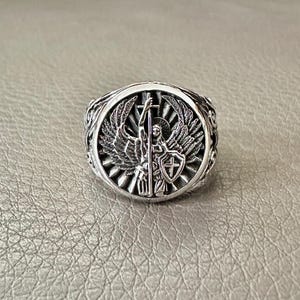 May include: A silver ring featuring an angel holding a cross and a shield with a cross. The angel has wings and is standing on a sunburst.