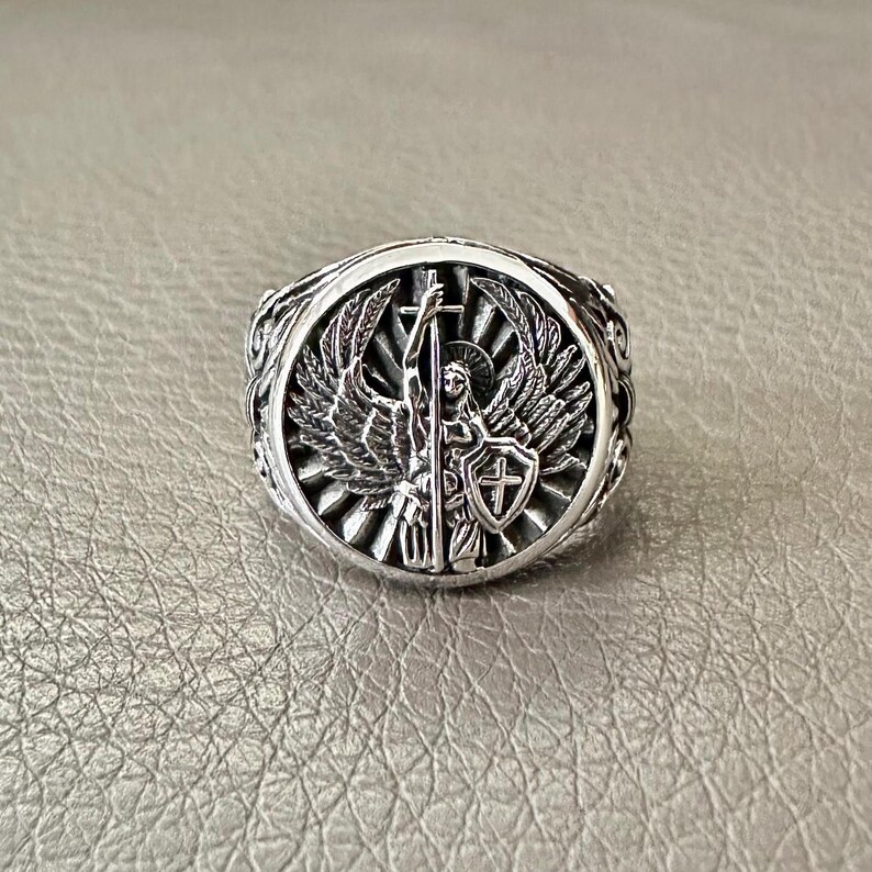May include: A silver ring featuring an angel holding a cross and a shield with a cross. The angel has wings and is standing on a sunburst.
