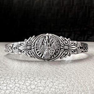 Könnte beinhalten: Silberfarbenes Armreif-Armband mit aufwendigem Design. Das Zentrum zeigt ein rundes Emblem mit einer geflügelten Figur, die einen Stab und einen Schild hält. Das Armband hat detaillierte Blumen- und Knotenmuster.
