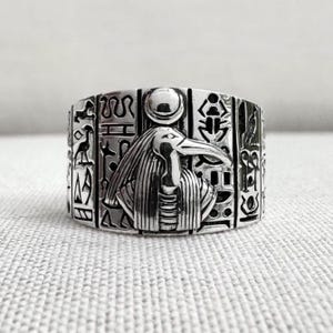 May include: A silver ring with an Egyptian theme. The ring displays a bird-headed deity at its centre, framed by hieroglyphs and other ancient symbols. The band is wide, with intricate engraved detailing, reflecting a historical aesthetic.