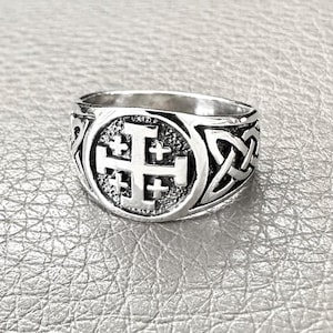 以下が含まれることがあります： 中央にエルサレム十字のデザインが施されたシルバーカラーのリング。リングはテクスチャード加工された背景と、側面にケルトノットのディテールが施されています。シグネットスタイルリングです。
