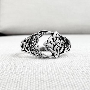 May include: A silver ring with an open design featuring Celtic knot patterns. The ring has intricate interwoven knotwork, with a prominent triquetra knot at the front. The ring is displayed on a white textured surface.