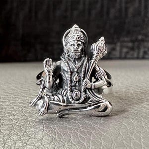 Peut inclure: Une bague en argent représentant une sculpture détaillée de la divinité hindoue Hanuman en position assise. La bague est en métal et a une finition texturée.