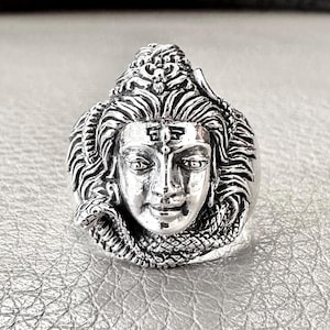May include: A silver ring featuring a detailed depiction of the Hindu deity Shiva, with a snake wrapped around his head.