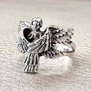 May include: A silver ring with a detailed angel figurine. The angel has outstretched wings and folded hands, with intricate feather detailing. The ring has a polished finish. The design is highly detailed.