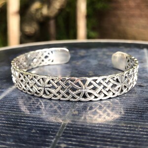 May include: Silver Celtic knotwork bangle bracelet. The bracelet is made of a thick, silver band with a detailed Celtic knot design.