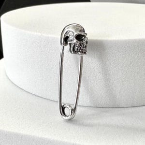 Puede incluir: Un imperdible plateado con una cabeza en forma de calavera. La calavera tiene detalles como cuencas de los ojos, cavidad nasal y dientes. El imperdible tiene un diseño clásico, con un extremo redondeado y un cierre.