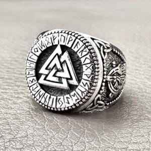 Puede incluir: Anillo plateado con un símbolo Valknut central rodeado de runas. El anillo presenta grabados laterales detallados y una banda texturizada, mostrando un diseño vikingo o nórdico.