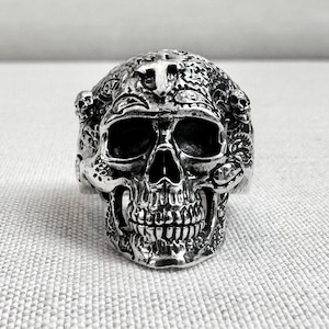 May include: A detailed silver skull ring with intricate carvings and a cross on the forehead. The ring features a full skull design with visible teeth and eye sockets, showcasing a gothic or biker aesthetic.