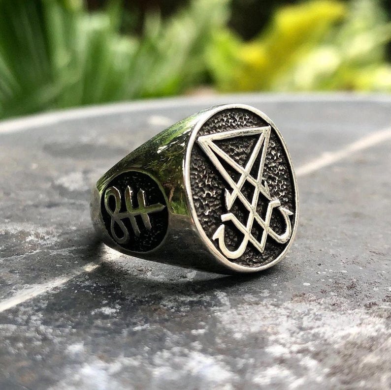 May include: A silver ring with a black sigil of a pentagram with an upside-down cross inside. The ring has a textured surface and a raised edge.