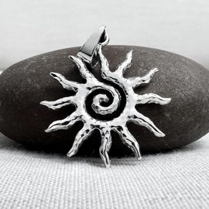 Könnte beinhalten: Silberfarbener Anhänger in Form einer Sonne mit einem spiralförmigen Zentrum. Die Sonne hat spitze Strahlen. Der Anhänger wird auf einem dunkelgrauen Stein präsentiert.