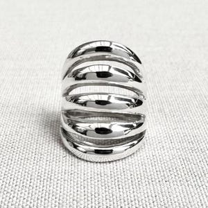 May include: A wide, polished silver ring featuring a multi-band design composed of five distinct, smooth, rounded horizontal bars that are connected at the sides, creating a series of open, parallel gaps between each metallic band.